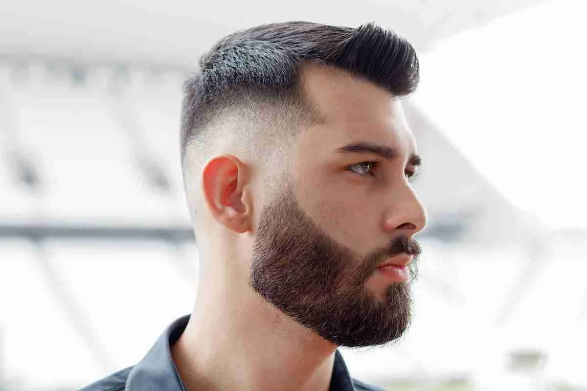Homem de barba cheia com undercut, lateral curta uniforme e topo penteado para trás com volume