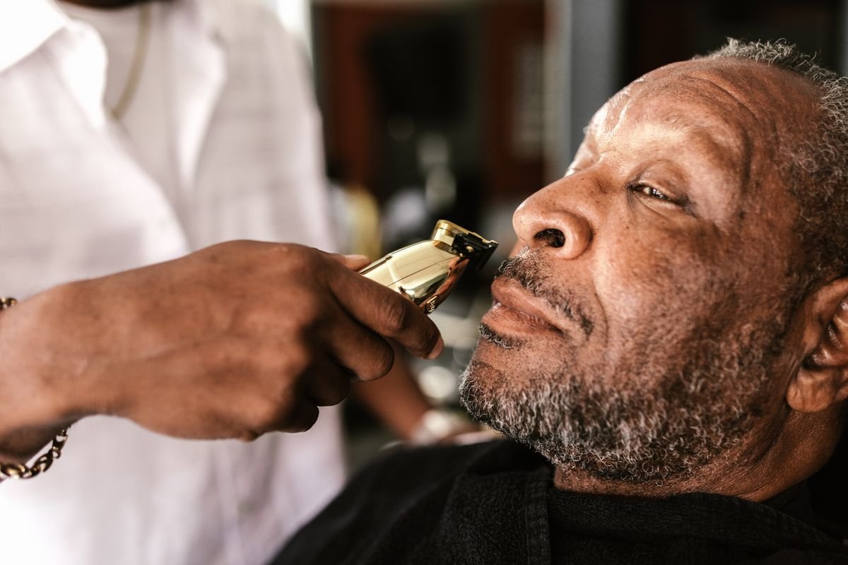 Barbeiro acertando com aparador a barba grisalha de um cliente mais velho
