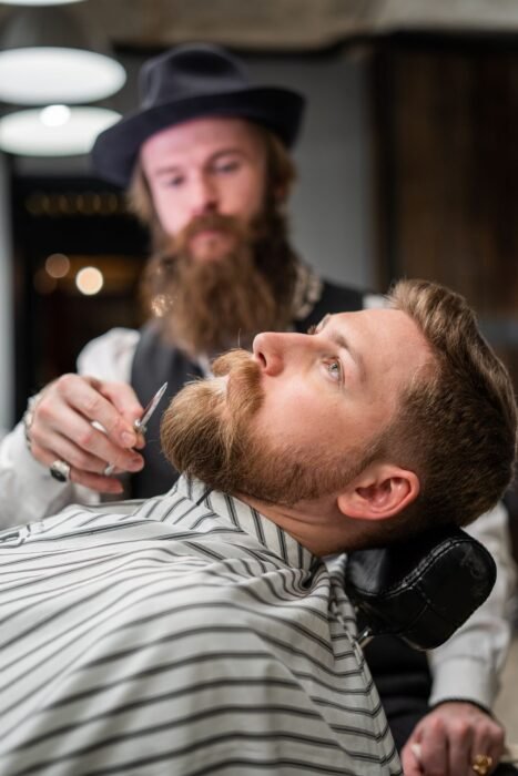 Barbeiro de chapéu ajustando a barba de um cliente na cadeira da barbearia