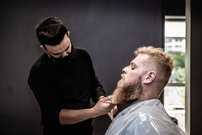 Barbeiro aparando a barba de um cliente com pente e tesoura