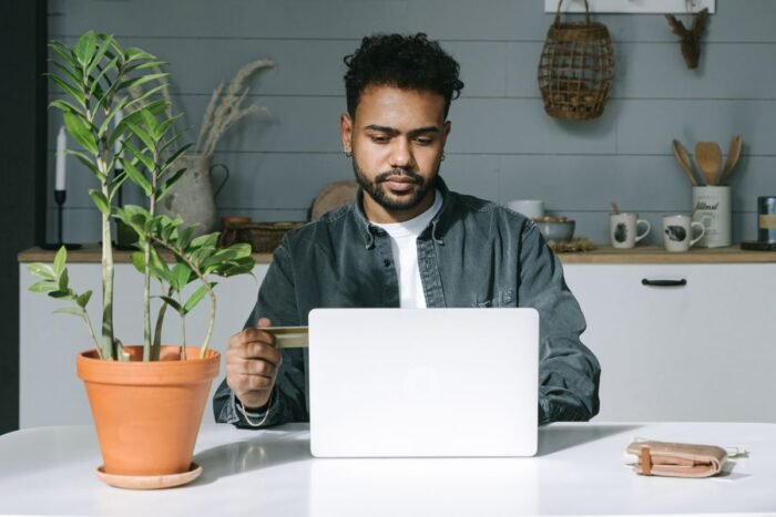 Homem de barba conferindo uma compra online no notebook com o cartão na mão
