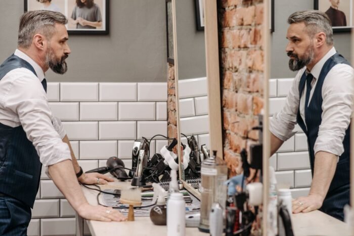 Homem de barba grisalha se olhando no espelho de uma barbearia