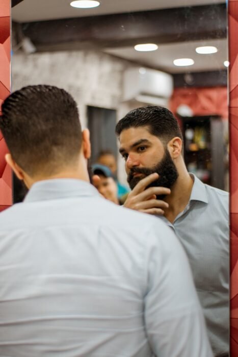 Homem de barba examinando o rosto no espelho da barbearia