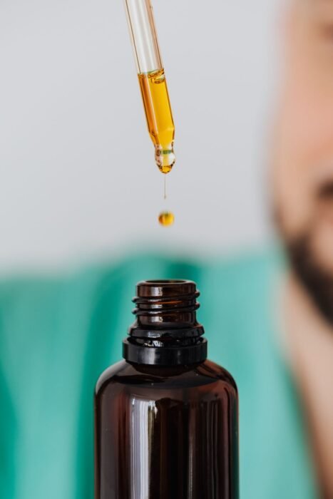 Conta-gotas pingando óleo dourado em frasco âmbar, com homem de barba desfocado ao fundo