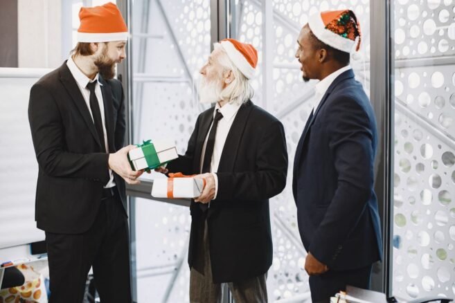 Três homens de terno e gorro natalino trocando presentes de amigo secreto no escritório